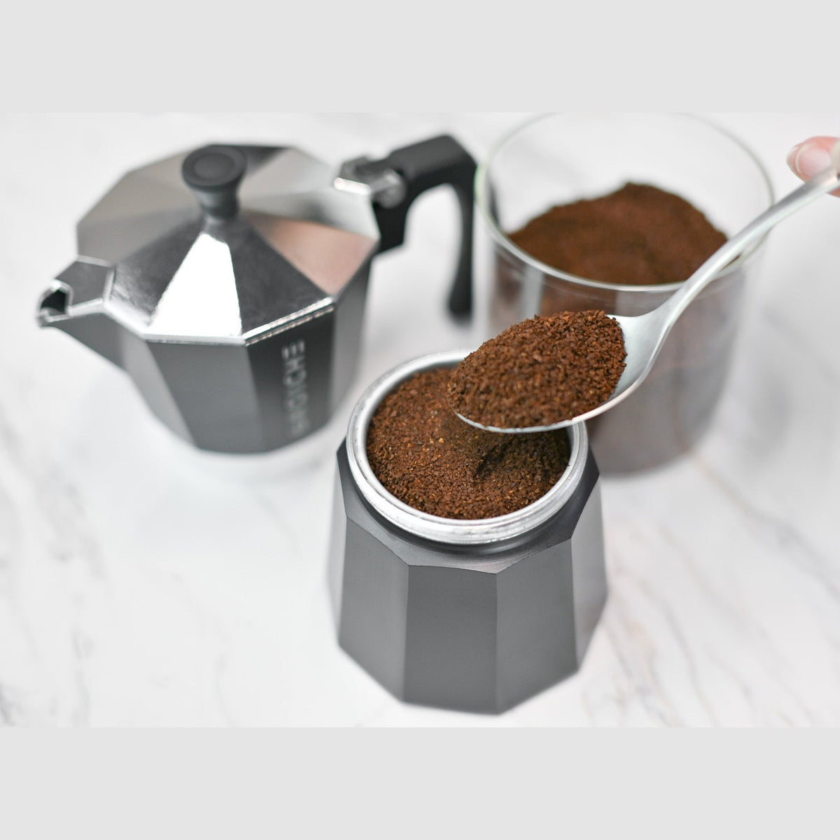 Dark grey moka pot filter basket filled with ground coffee being scooped by spoon