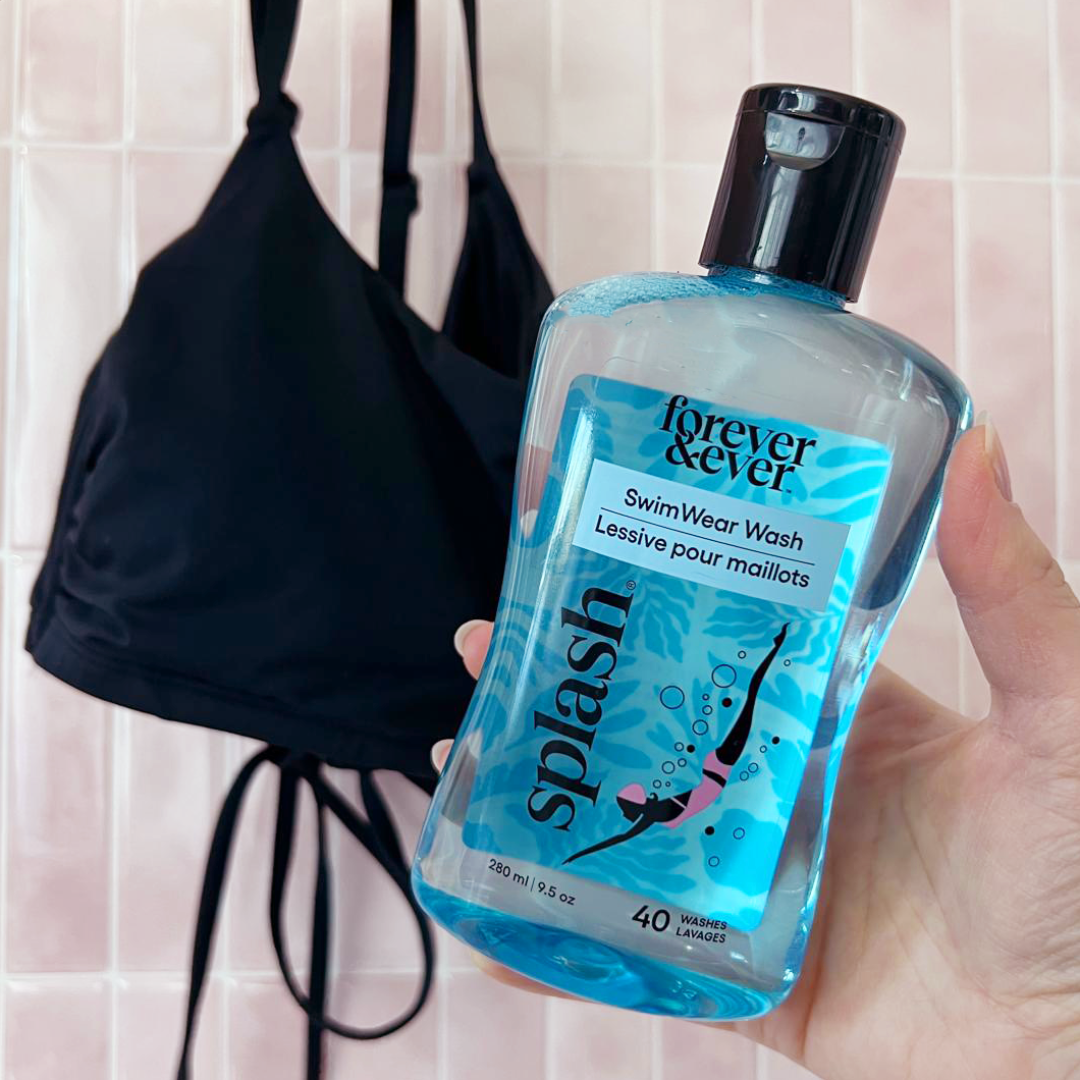 A hand holds Forever & Ever Splash® Swimwear Wash before a black bikini top on a pink tiled wall.
