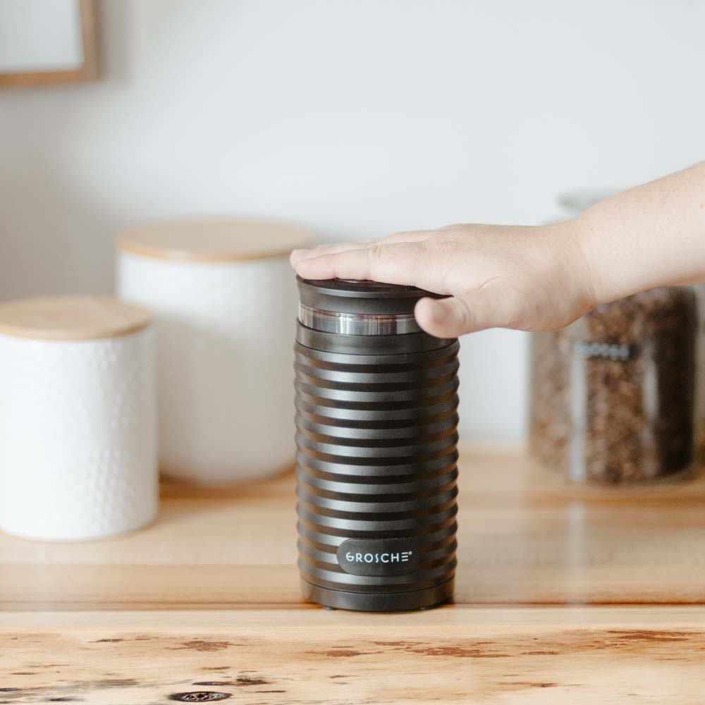 Hand pressing top lid of black ribbed Grosche electric blade coffee grinder demonstrating use