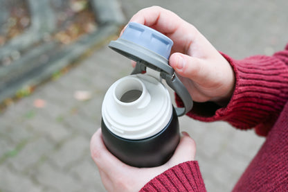 A person in a red sweater opens a Grosche Kamloops 24oz Insulated Chug Lid Water Bottle outdoors.