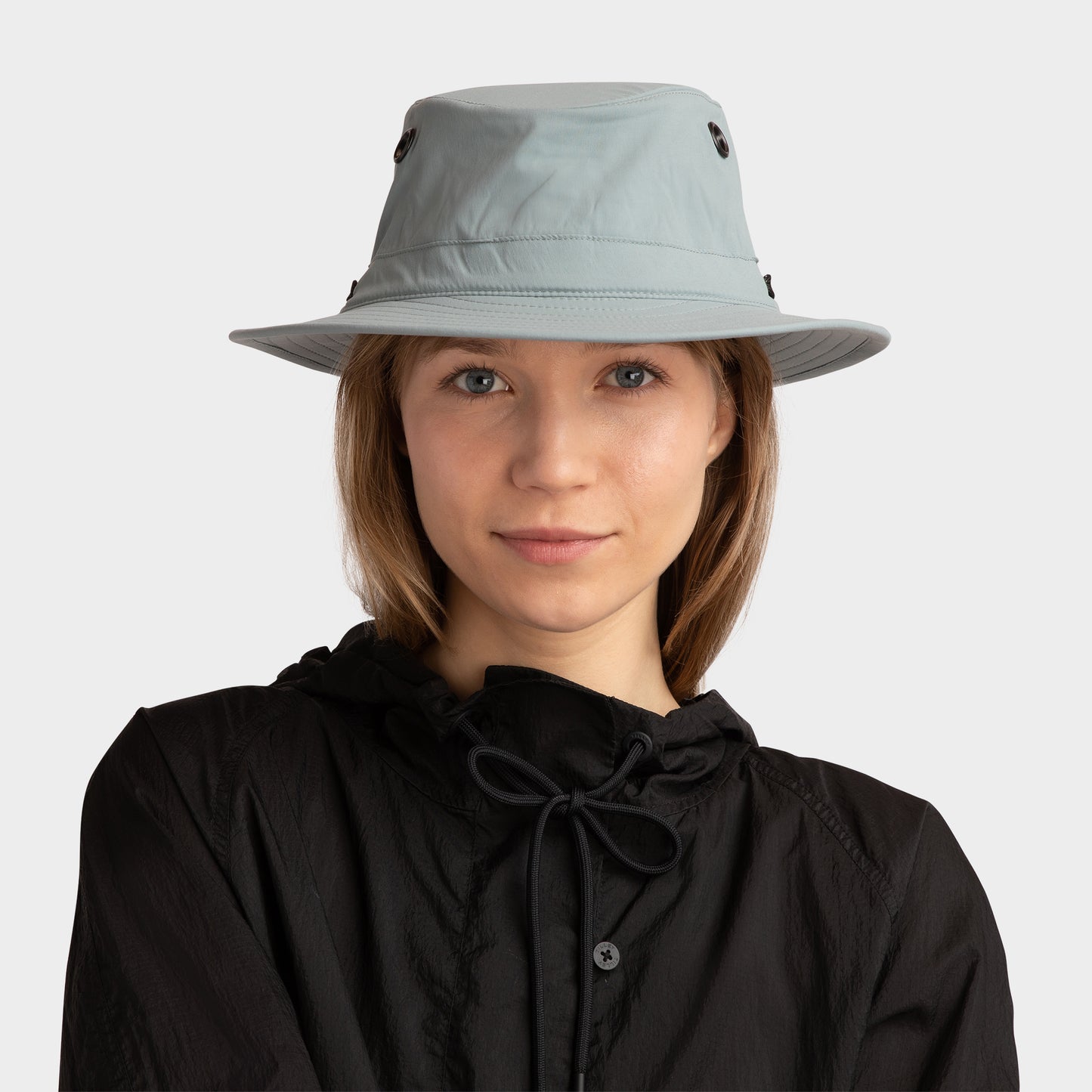 A woman wears a Tilley T5 Adventure Ultralight Hat and black jacket, facing forward against a plain background.