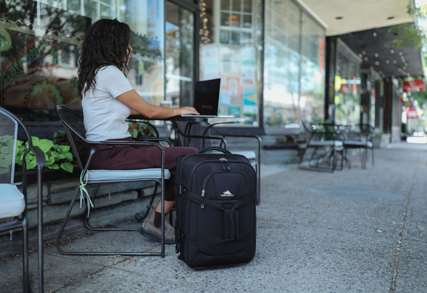 Woman sitting at a small table outside working on a laptop with the High Sierra Pendall carry-on beside her.
