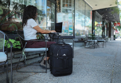 Woman sitting at a small table outside working on a laptop with the High Sierra Pendall carry-on beside her.