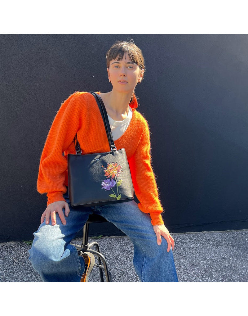 Woman wearing blue jeans and a bright orange sweater, sitting on a stool outside with the Espe Glee Backpack converted with shoulder straps, slung on one shoulder