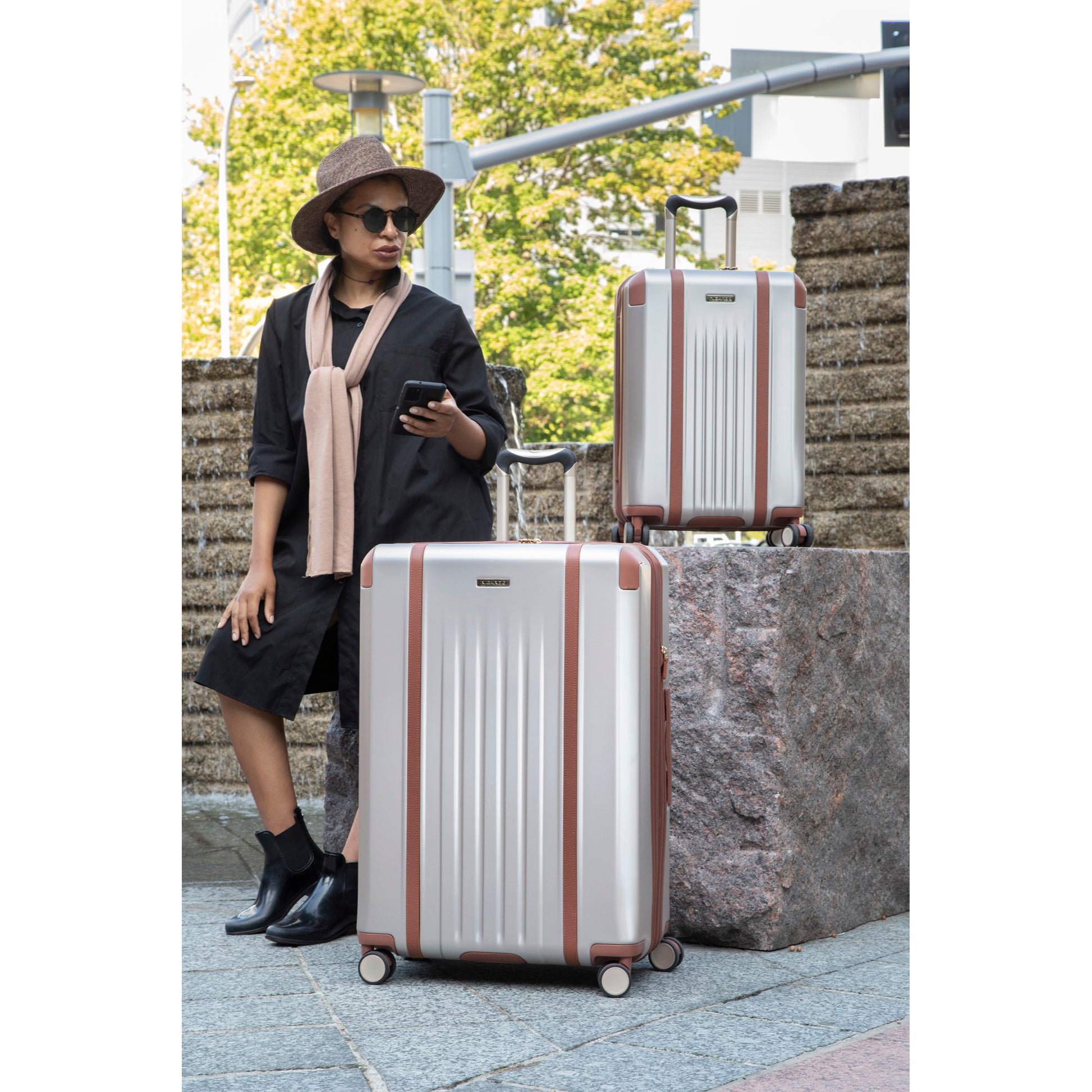 Woman standing near brick wall with champagne Montecito 2.0 large and carry-on suitcases beside her.