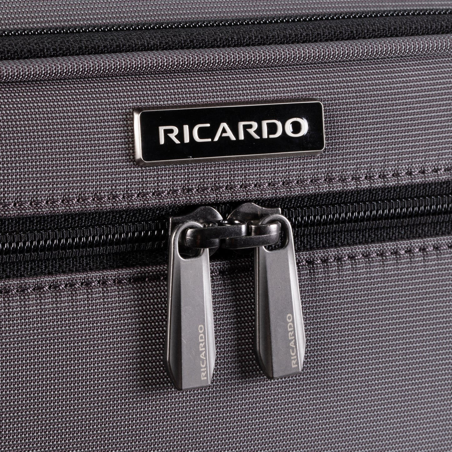 Close-up of Ricardo Beverly Hills Montecito 3.0 Softside Carry-on Spinner in gray with double zippers and logo above.