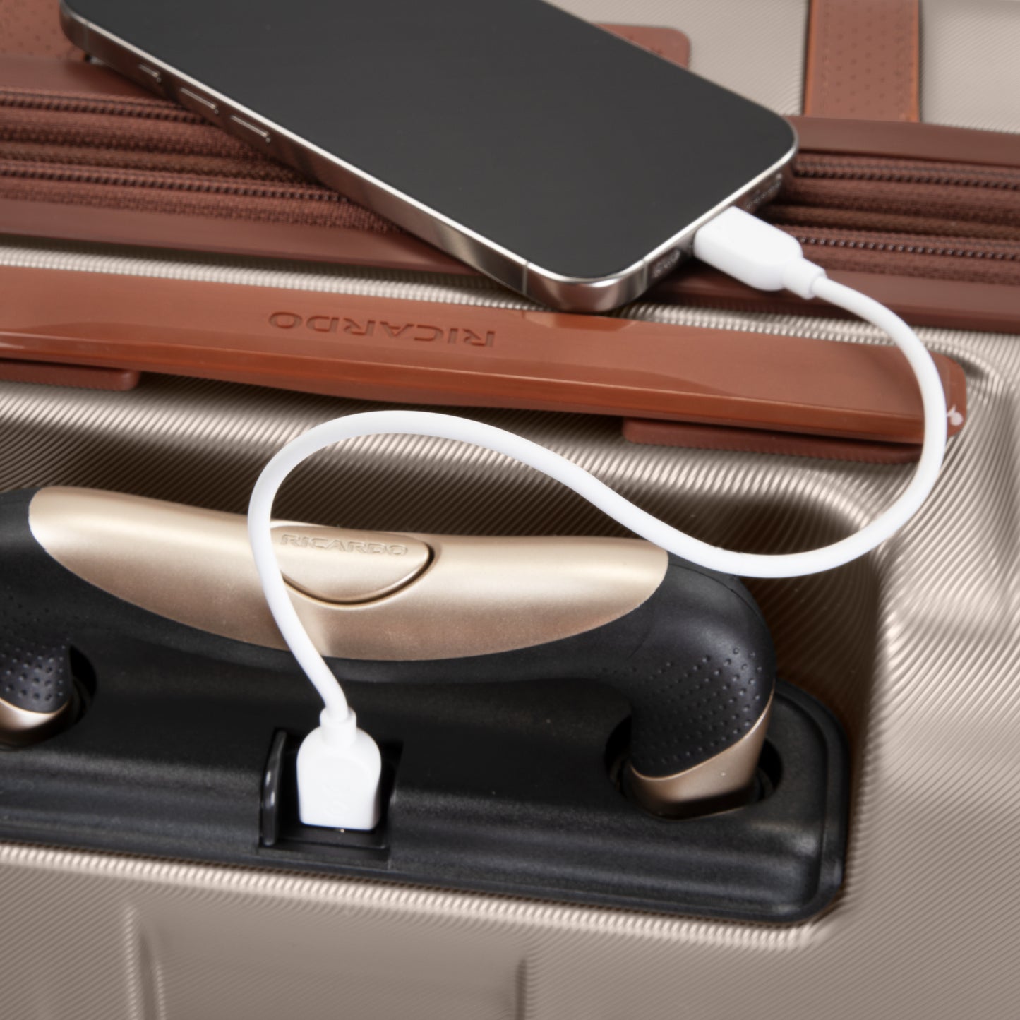 A smartphone charges via cable on the Ricardo Beverly Hills Montecito 2.0 carry-on with built-in USB port.