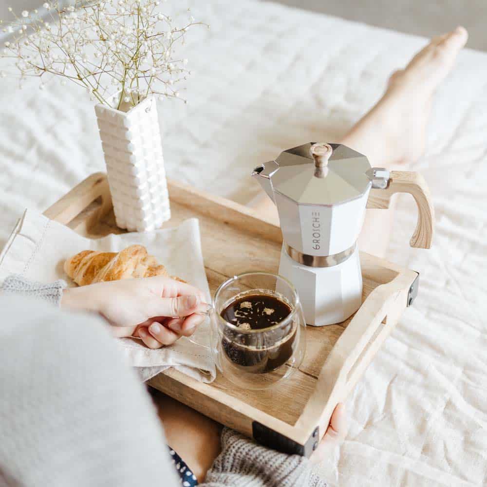 White moka pot with wooden handle on breakfast tray with croissant and glass cup