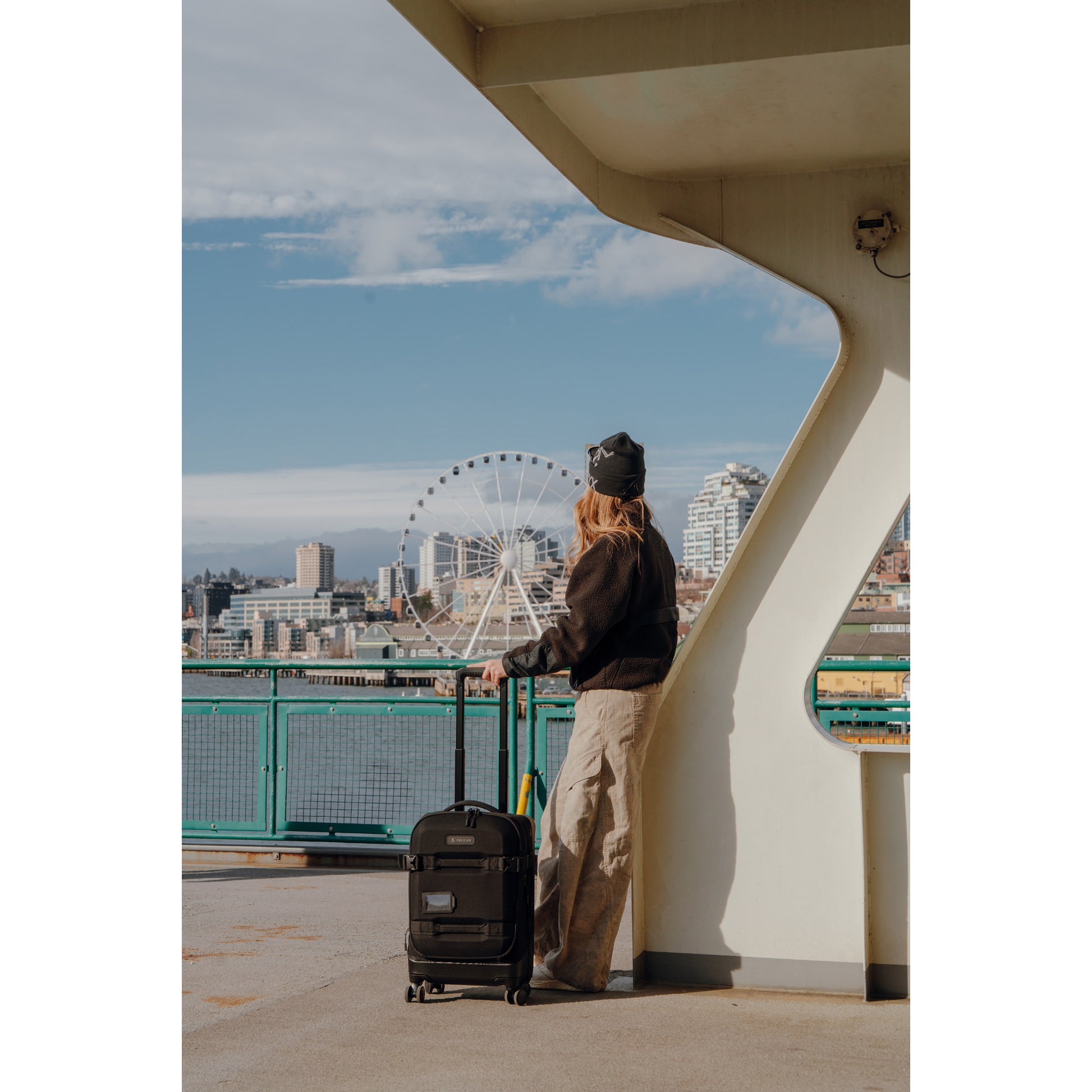 Traveler with a Pelican™ Aegis Carry-on Duffel stands on a ferry, gazing at the city skyline and Ferris wheel.