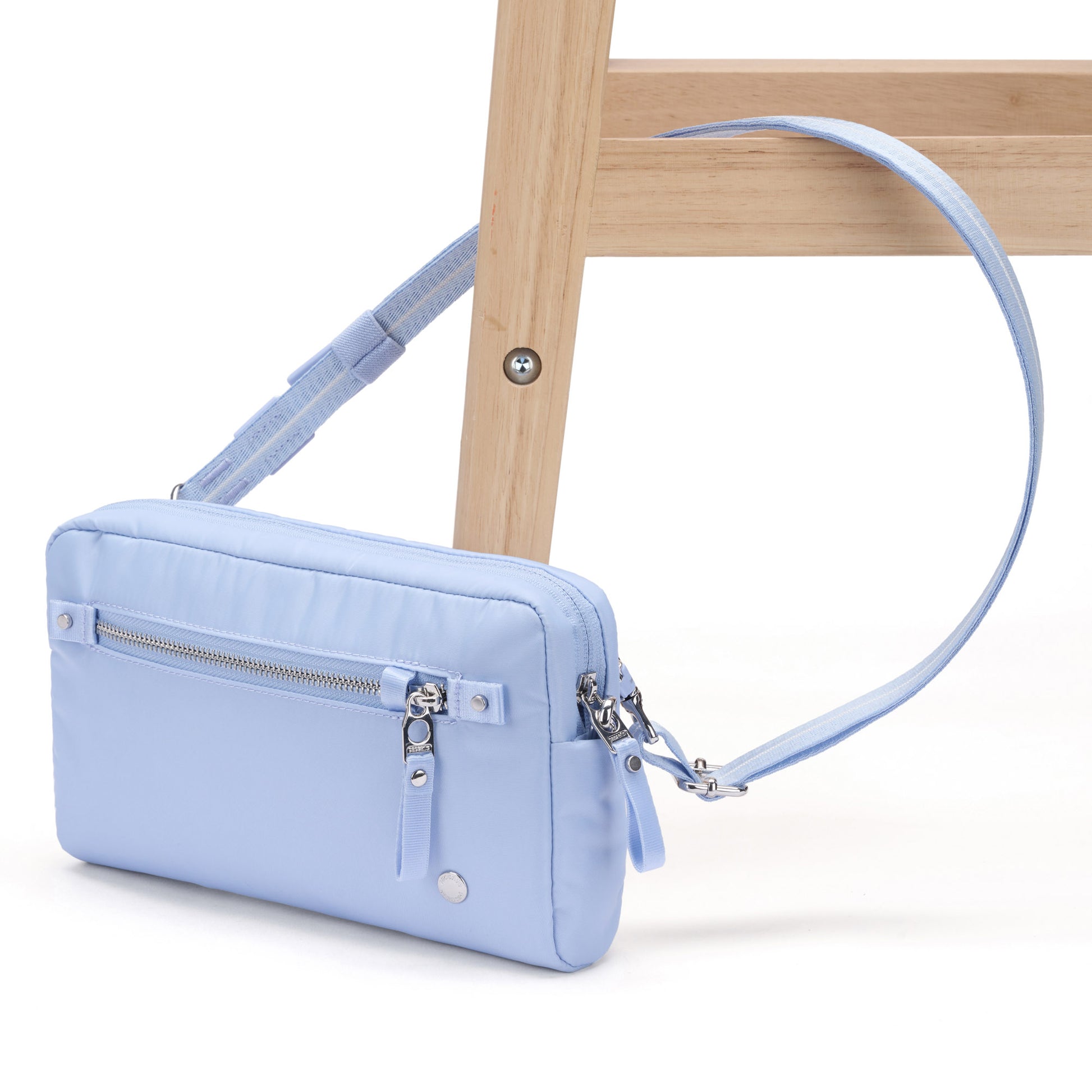 The Pacsafe® W Anti-theft 3-in-1 Sling by Pacsafe hangs from a wooden chair leg, light blue with silver zippers.