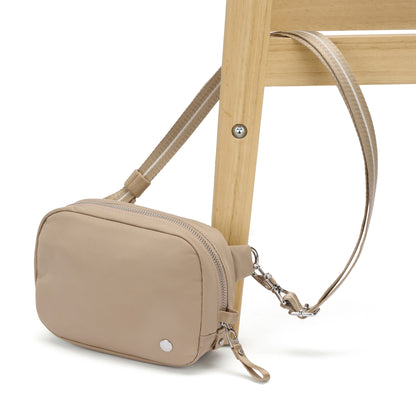 The Pacsafe® W Anti-theft Micro Sling in beige recycled polyester is strapped to a wooden chair leg.
