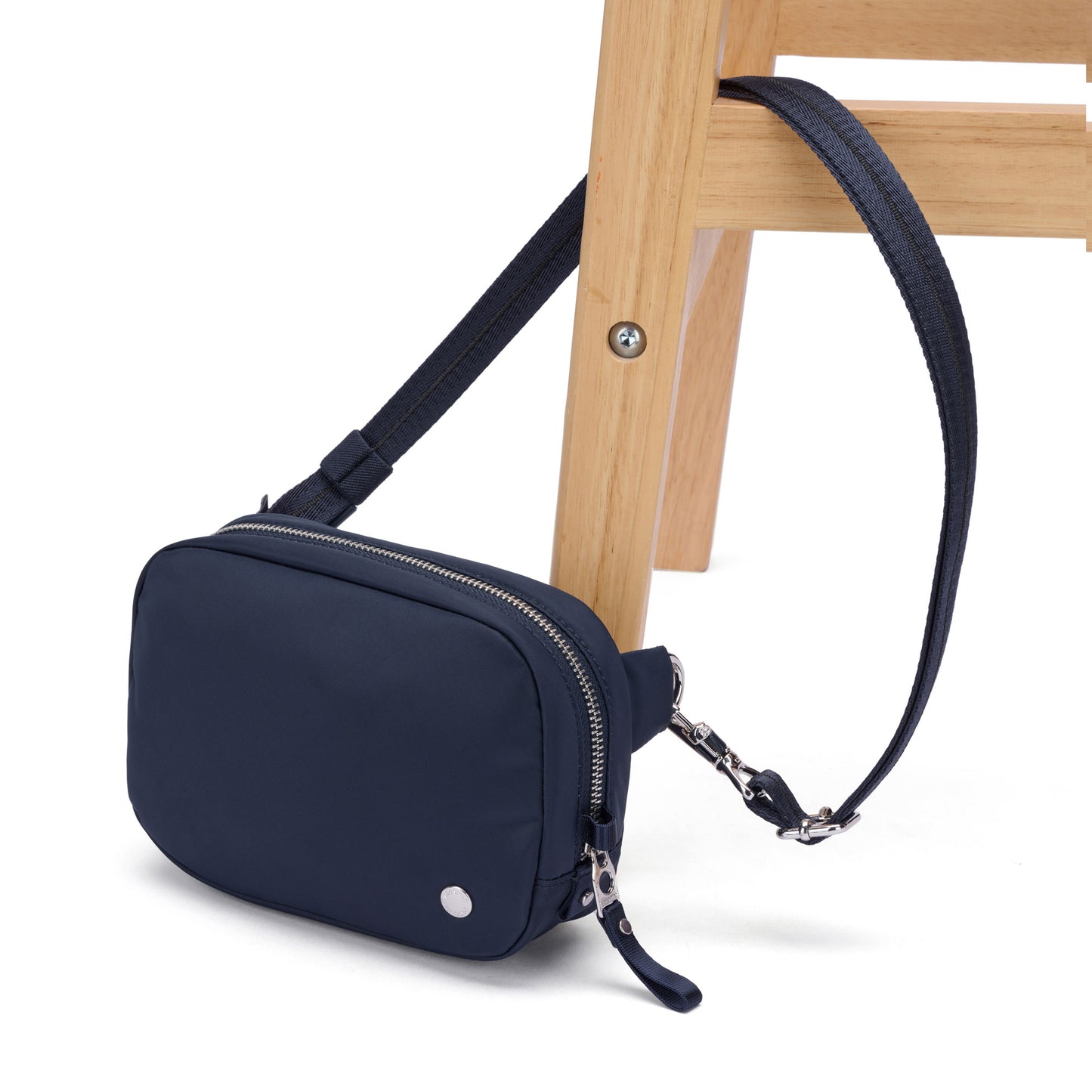 The Pacsafe® W Anti-theft Micro Sling in navy blue rests on the leg of a light wooden chair.