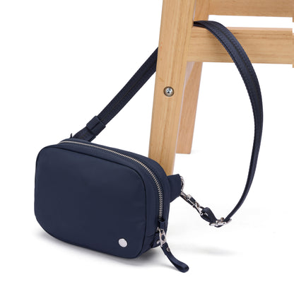 The Pacsafe® W Anti-theft Micro Sling in navy blue rests on the leg of a light wooden chair.