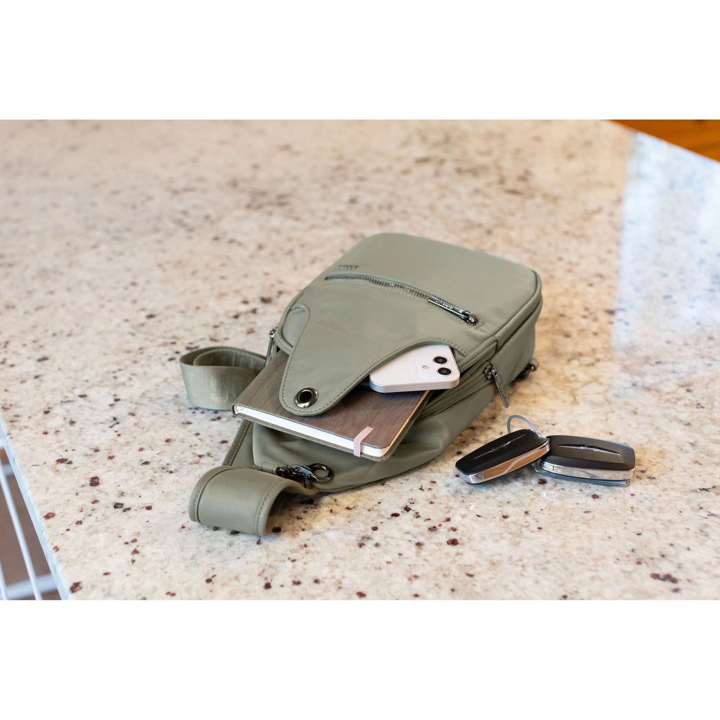 Lifestyle image of the Roots Travel Sling Bag in sage colour, laying on a marble countertop with a notebook and phone sticking out and car keys beside it.