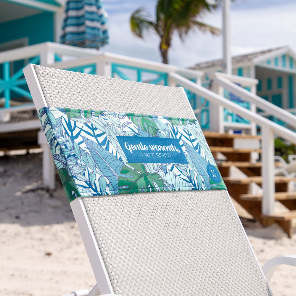 K5 Reserved Chair Sleeve by K5 on a white lounge chair, with a beach accessory, set near turquoise cabins on sand.