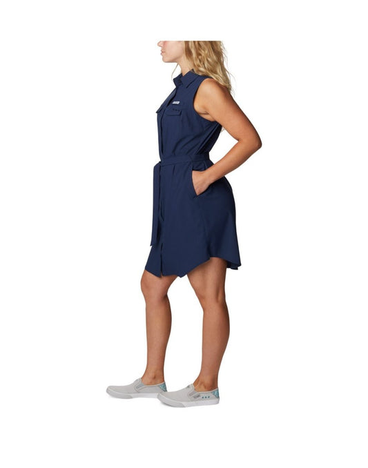 Product Image – Woman wearing light grey running shoes and Columbia Women's PFG Sun Drifter™ Woven Dress II in collegiate navy, side view with hands in pockets