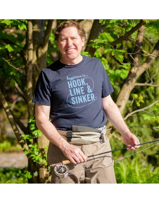 Product Image – Man wearing khaki pants and Happiness Is... Men's Hook, Line & Sinker T-Shirt  in navy holding a fishing rod with trees in the background