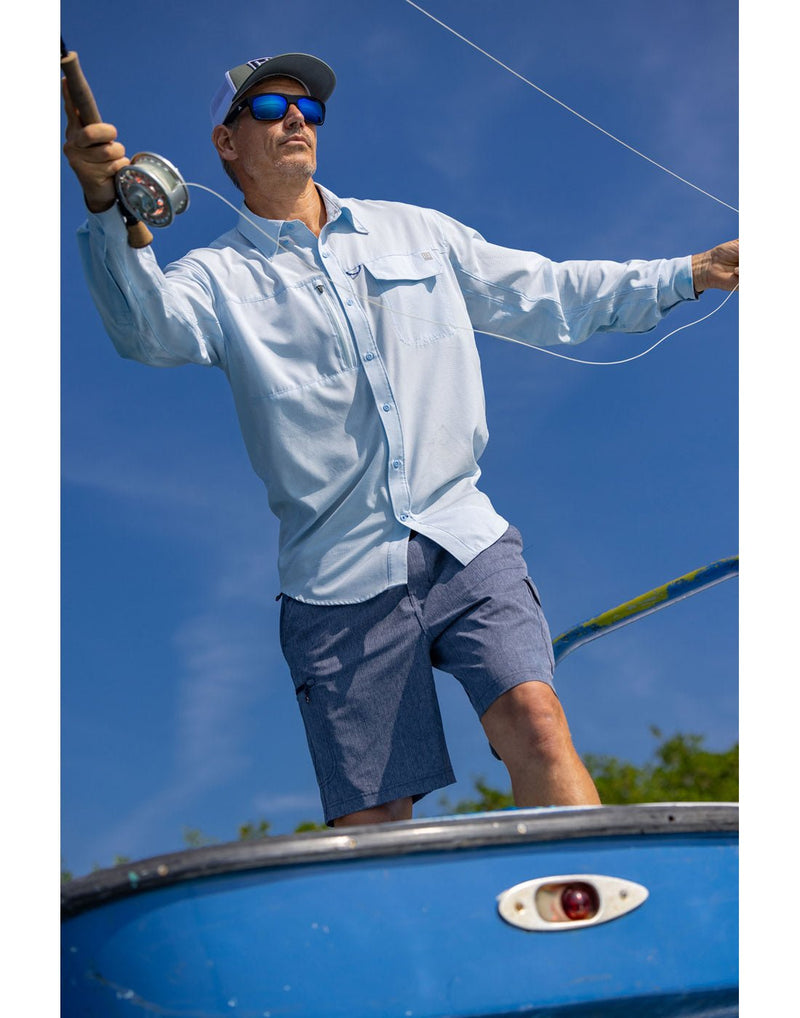 Lifestyle image, Front view of a man wearing a Huk Men's A1A Button-Down Shirt in Ice Water blue colour and blue shorts.  Standing on a boat and fishing.