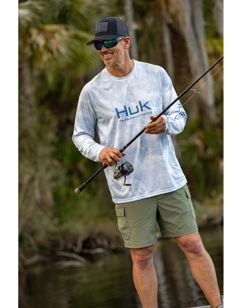 Lifestyle front view of a man wearing the Huk Men's Mossy Oak Pursuit Performance Shirt in Mossy Oak Stormwater Bonefish colour, green cargo shorts and holding a fishing pole.