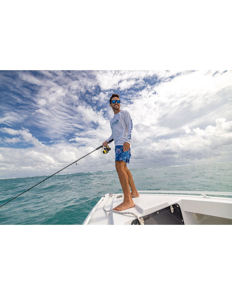 Lifestyle image, front left side view of a man standing on the bow of a boat wearing the Huk Men's Pursuit Performance Shirt in white, blue patterned shorts and holding a fishing rod.
