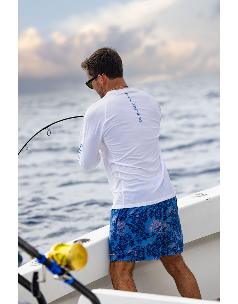 Lifestyle image, back view of a man wearing   the Huk Men's  Pursuit Performance Shirt in white  and blue patterned shorts and holding a fishing rod.