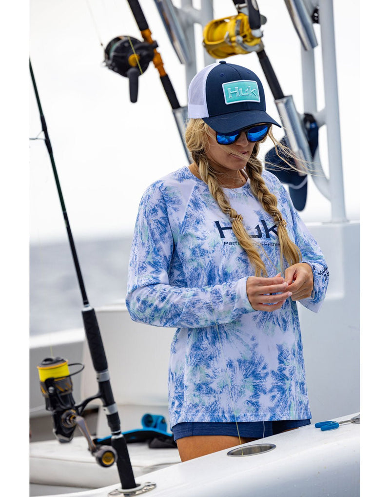 Lifestyle image of a woman wearing the Huk Women's Pursuit Performance Shirt in Aqua Dye White colour, blue shorts and a Huk logoed baseball style hat.