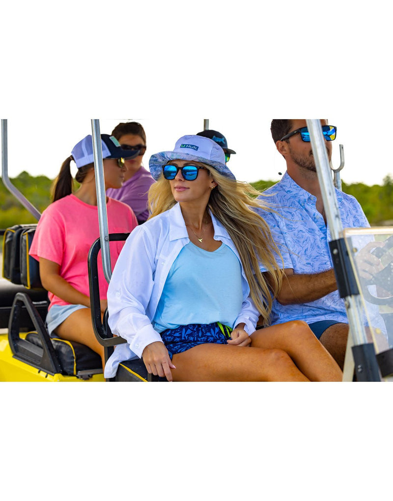 Lifestyle image of woman wearing Huk Women's Tide Point Button-Down Shirt in white, open on top of blue t-shirt, riding on a golf cart with four other people all wearing sunglasses