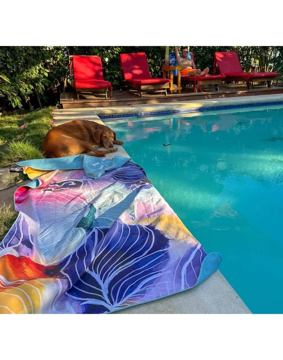 Lifestyle image of abstract flower K5 Microfibre Beach Towel, laid on the ground beside a pool, with poolside chairs and dog visible in the background.