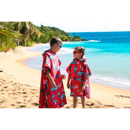 Two kids in red K5 Microfibre Ponchos and sunglasses stand on a sandy beach by the ocean.