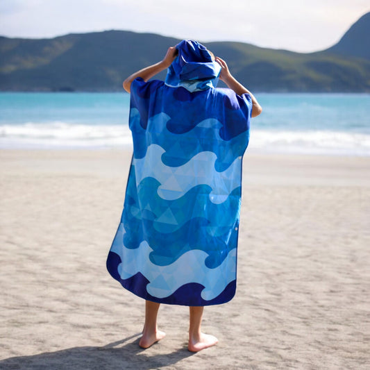 Product Image – Teen in a blue K5 Microfibre Poncho stands on a sandy beach facing the sea and distant hills.