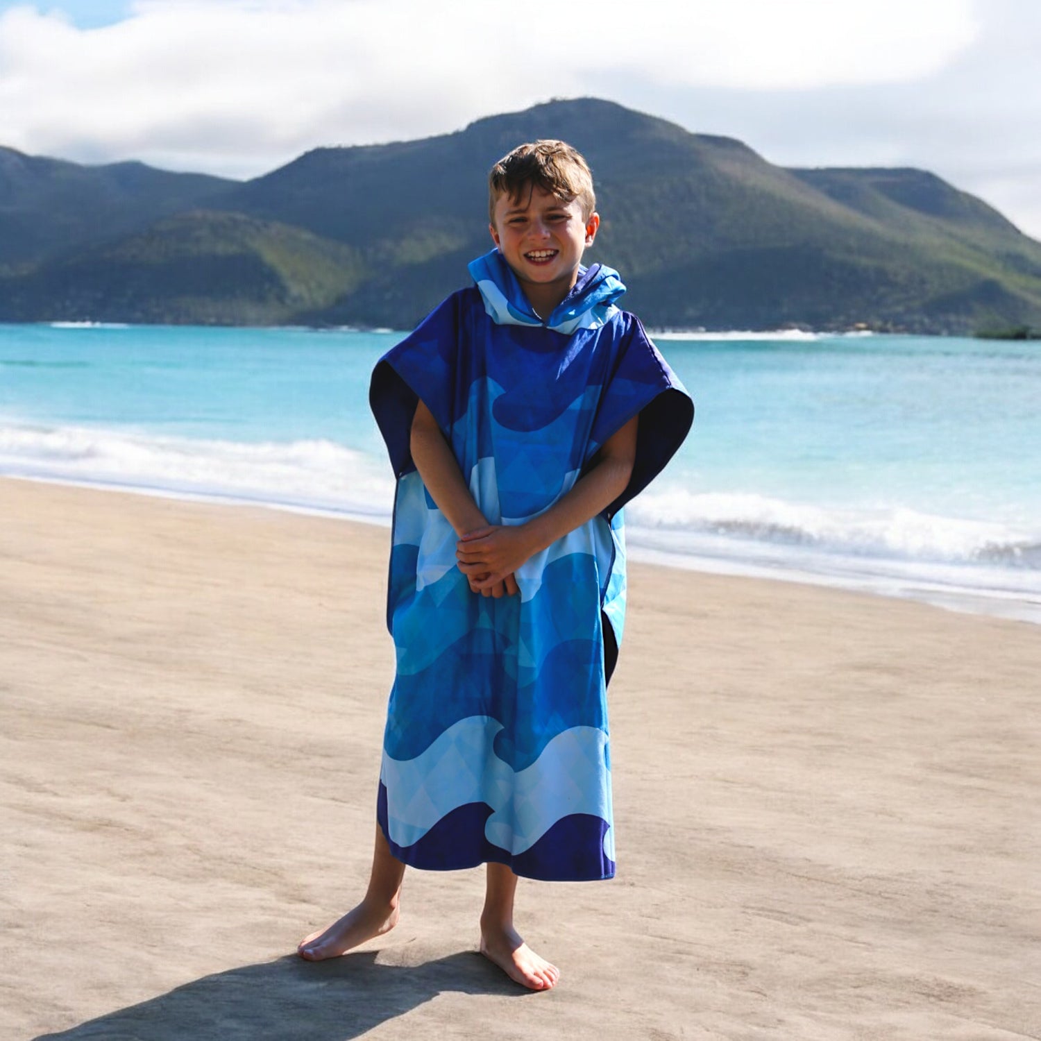 Smiling boy in a K5 Microfibre Poncho - Teen stands on shore with mountains and ocean behind.