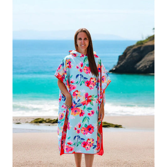 Product Image – Woman wearing the K5 Microfibre Poncho stands on a sandy beach with turquoise water and rocky cliffs behind.