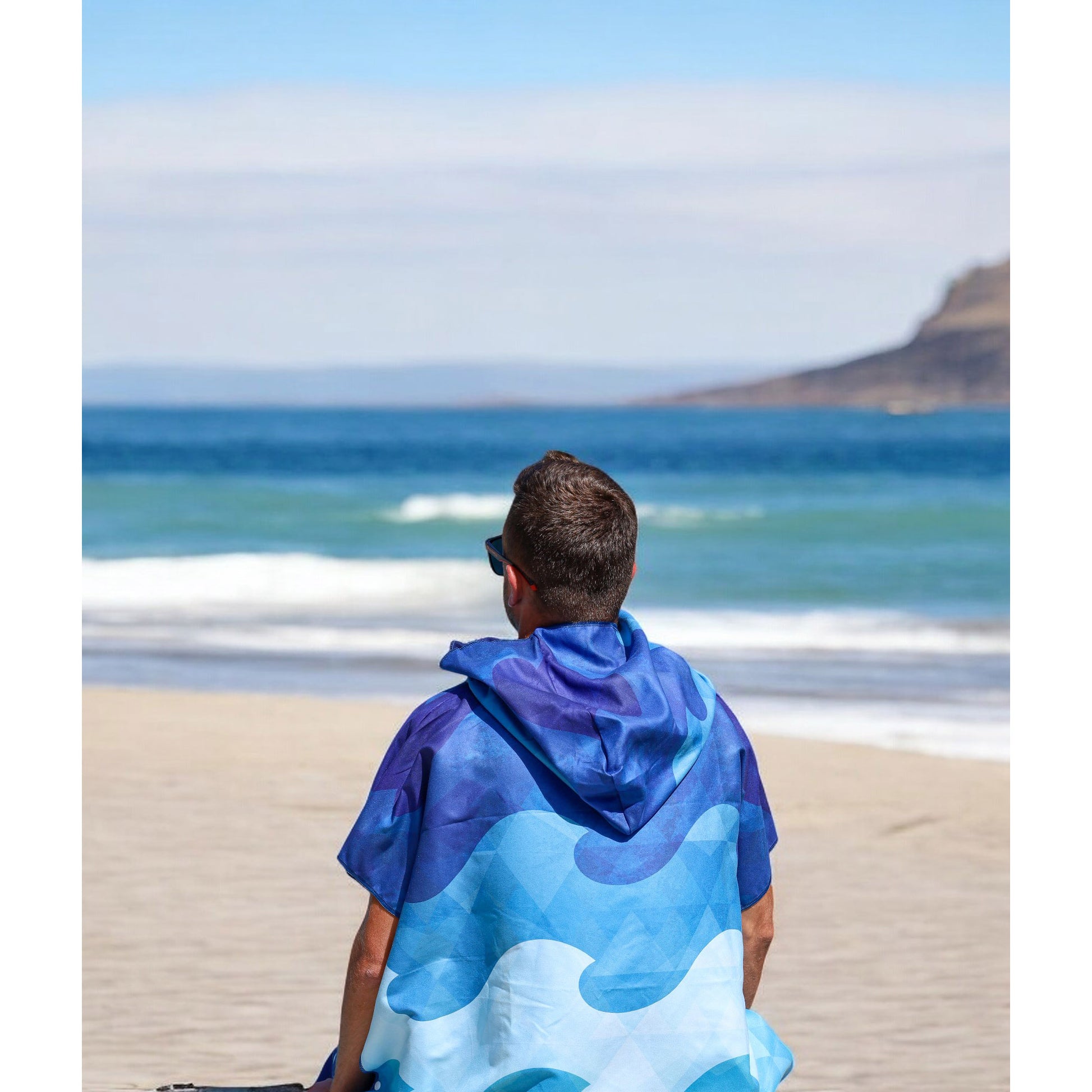 Someone wearing a K5 Microfibre Poncho sits on the beach, gazing at the ocean under a clear sky.