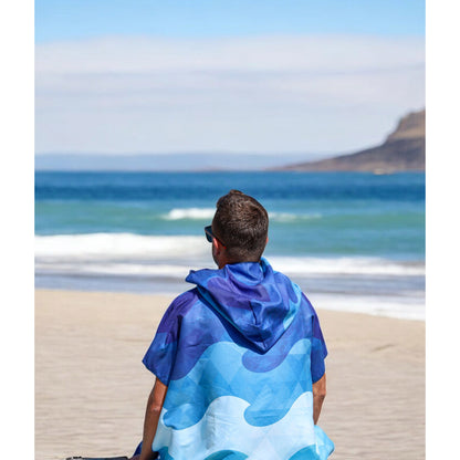 Someone wearing a K5 Microfibre Poncho sits on the beach, gazing at the ocean under a clear sky.