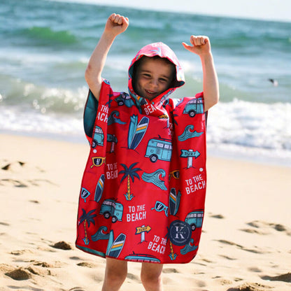 Child wearing K5 Microfibre Poncho - Kid stands on sandy shore, arms raised with ocean waves behind.
