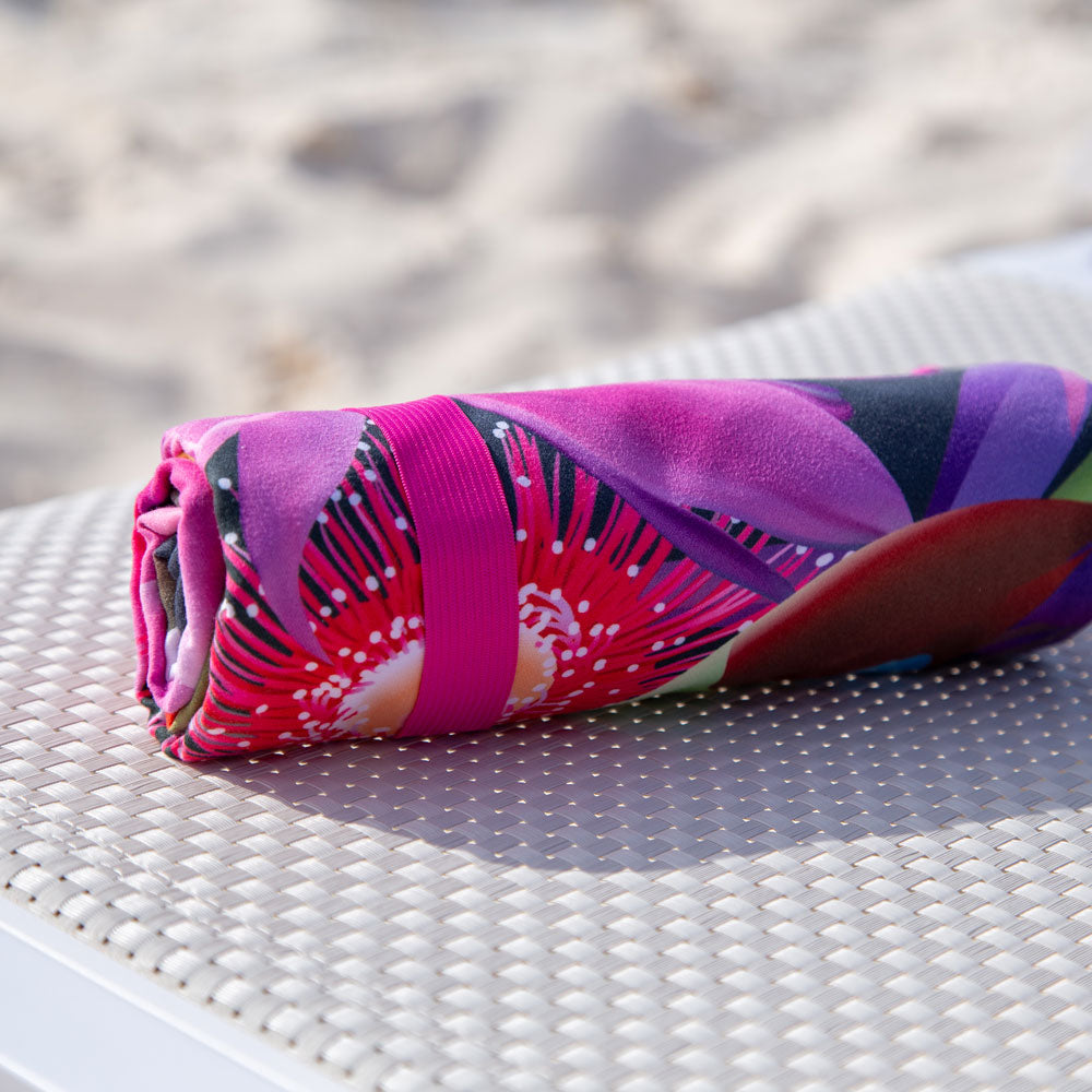 The K5 Microfibre Beach Towel, rolled up and colorful, sits on a white textured beach surface.