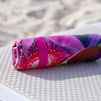 The K5 Microfibre Beach Towel, rolled up and colorful, sits on a white textured beach surface.