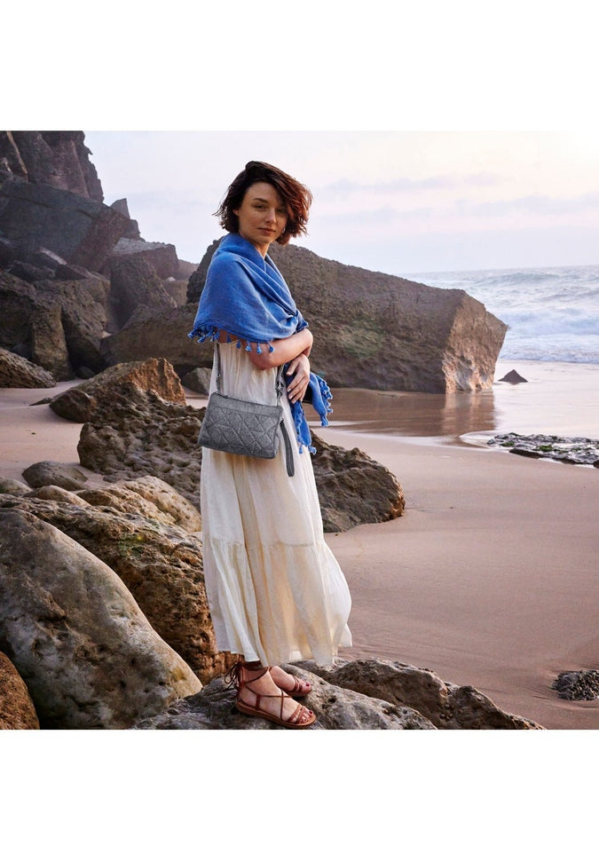 Lifestyle image of a woman at a rocky beach wearing the  view of the Travelon Boho Anti-Theft Clutch Crossbody in Grey.