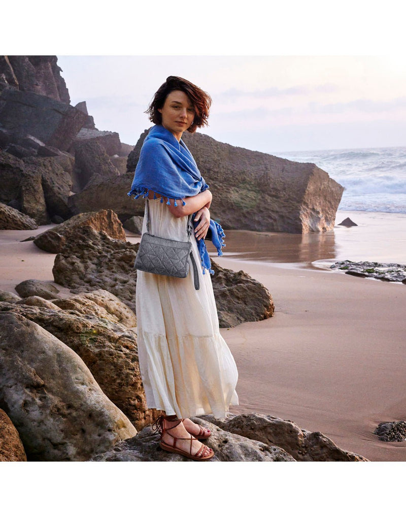 Lifestyle image of a woman at a rocky beach wearing the  view of the Travelon Boho Anti-Theft Clutch Crossbody in Grey.