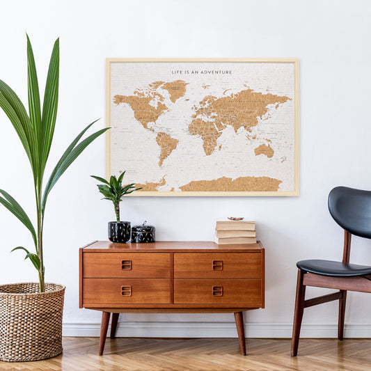 Product Image – Lifestyle image of Travel Board Large World Map hanging on a wall above a side table with books and a plant on it with a large plant and chair beside