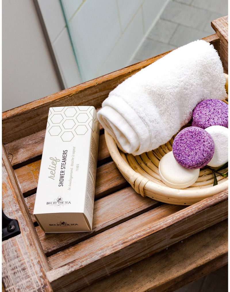 Two white and two purple Bee by the Sea shower steamers and a white rolled up towel on a bamboo basket with product box beside, both inside a wooden crate