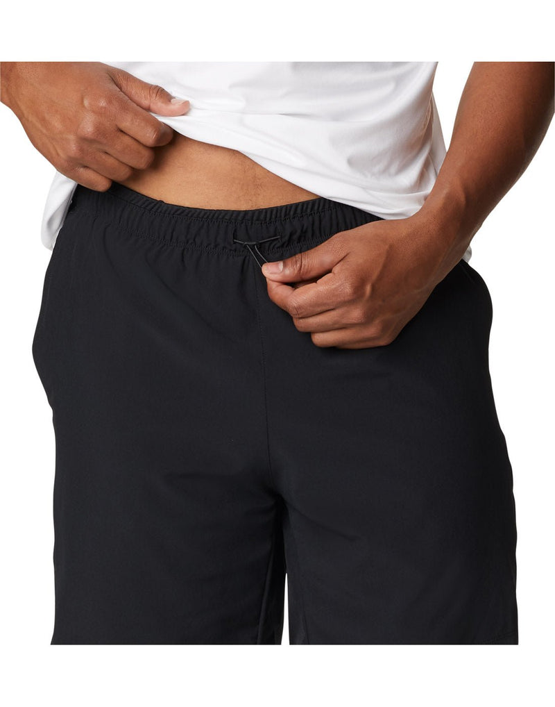 Close up of man wearing a white t-shirt and the Columbia Men's Alpine Chill™ Zero Shorts in black, front view, holding shirt up to show the drawstring at front centre of shorts