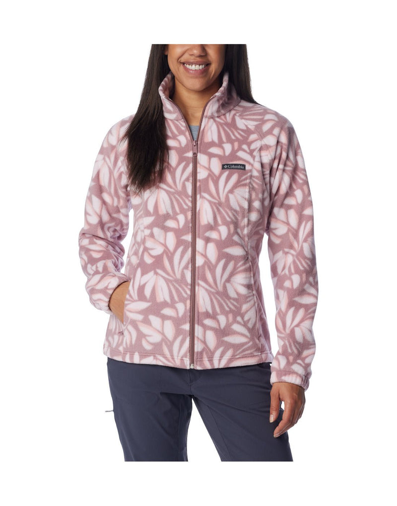 Woman wearing navy blue pants and Columbia Women's Benton Springs™ Printed Full Zip Fleece Jacket in fig areca, pink with white floral pattern, front view, zipped up with one hand in pocket