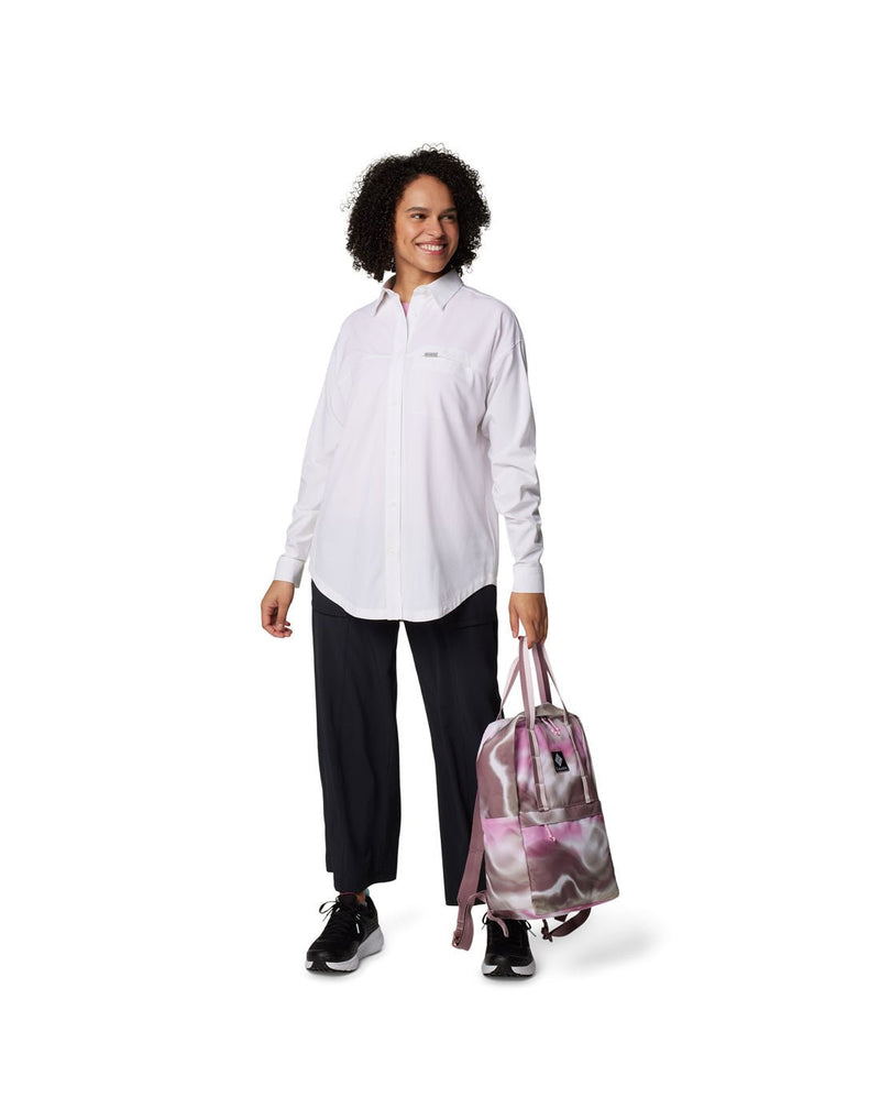 Woman wearing black pants, black running shoes, and Columbia Women's Boundless Trek™ Layering Long Sleeve Shirt in white, holding a backpack in one hand