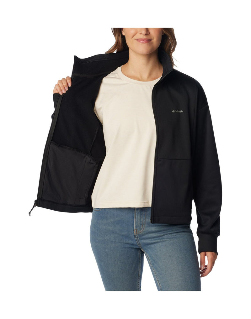 Woman wearing blue jeans and Columbia Women's Boundless Trek™ Tech Full Zip Jacket in black, unzipped, front view with white t-shirt underneath, holding one side of jacket open to show interior