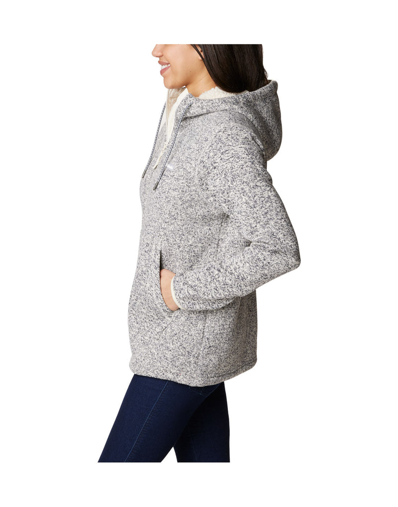 Left side view of a woman wearing the Columbia Women's Sweater Weather™ Sherpa Full Zip Hooded Jacket in Chalk Heather colour, hood down, hand in side pocket.
