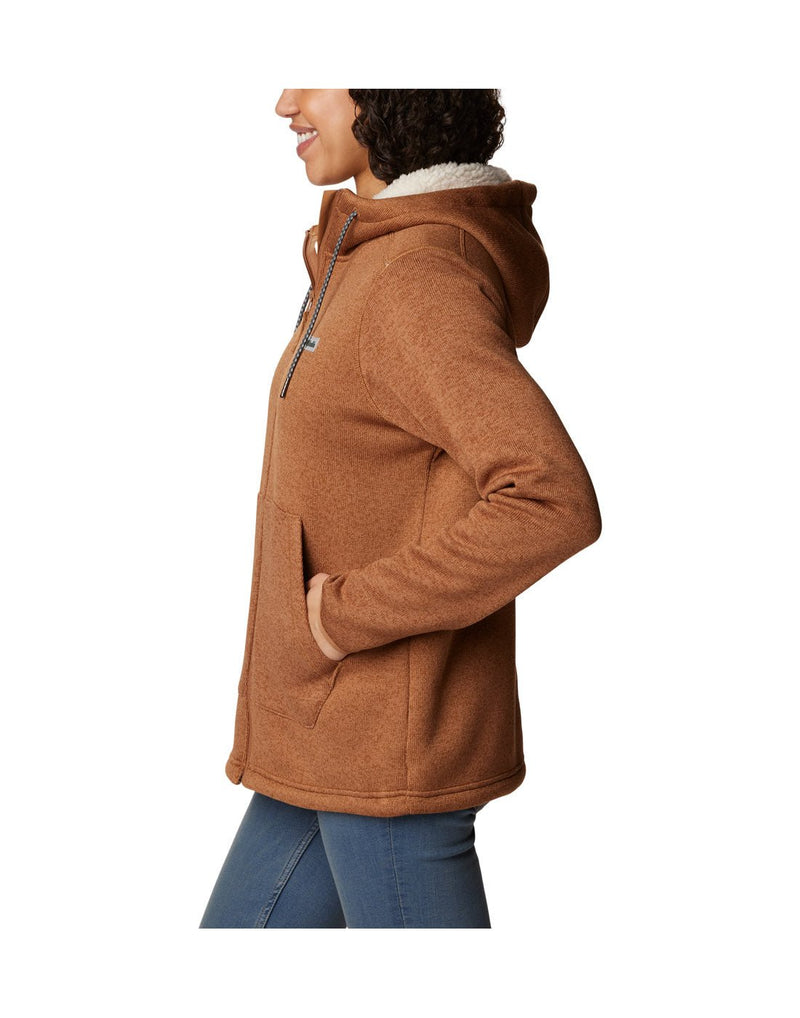 Left side view of a woman wearing the Columbia Women's Sweater Weather™ Sherpa Full Zip Hooded Jacket in Camel Brown Heather colour, hood down, hand in left side pocket.