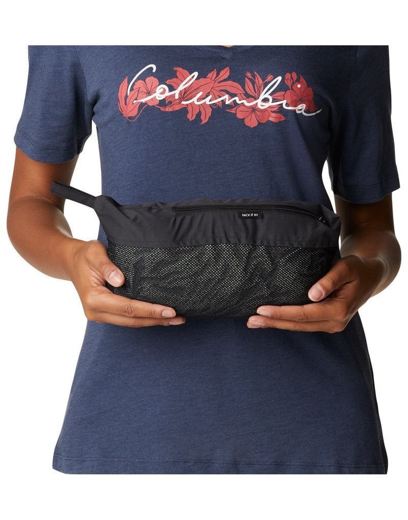 Woman wearing navy graphic t-shirt, holding Columbia Women's Weekend Adventure™ Long Shell Jacket in black, folded up into its attached mesh pouch