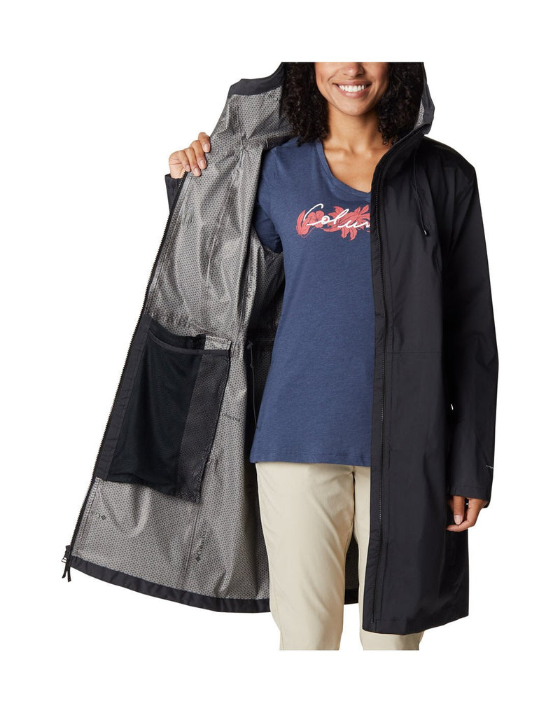 Woman wearing khaki pants, navy graphic shirt, and Columbia Women's Weekend Adventure™ Long Shell Jacket in black, unzipped, front view, holding one side of jacket open to show interior lining