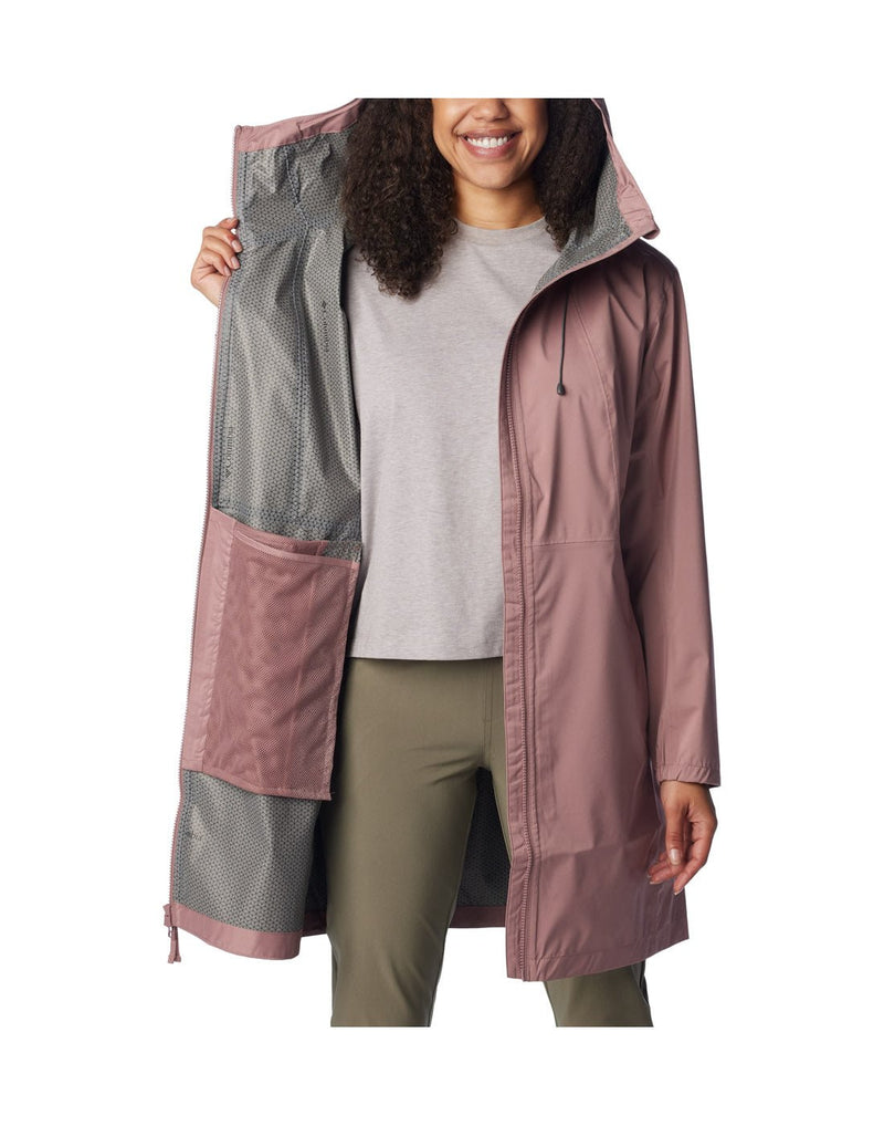 Woman wearing green pants, grey shirt, and Columbia Women's Weekend Adventure™ Long Shell Jacket in fig, unzipped, front view, holding one side of jacket open to show interior lining